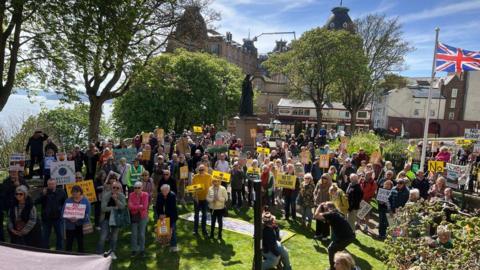 A group of people are holding signs, stood on a patch of grass surrounded by trees. There are civic building in the background with a clock tower.
