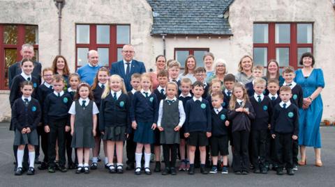Clann-sgoile bun-sgoile nan seasamh air an èideadh ann an deiseachan-sgoile. Tha triùir fhireannach agus seachdnar bhoireannach san dealbh.