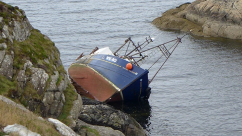 Bàta-iasgaich air a cliathaich san uisge agus i air na creagan. Tha an soitheach gorm agus donn.
