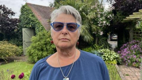 A woman with grey hair, dark glasses and a blue top with a silver necklace in a garden with bushes and purple flowers