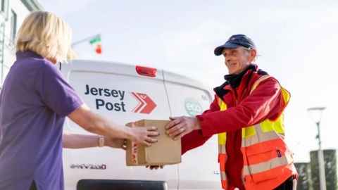 A woman with short blond hair wearing a purple polo shirt takes a small parcel from a postal worker dressed in red and a high-visibility jacket, standing in front of a white Jersey Post van.