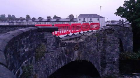 A row of five plastic fence segments have been erected along Nether Bridge. The wall of the stone bridge has been destroyed. A road of houses can be seen in the background.