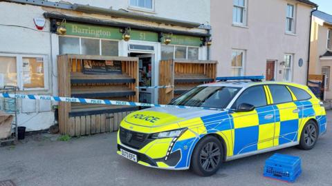 A police car is parked outside the village post office - a small shop with residential homes either side and on top of it. A police cordon is in place to stop people entering through the shops open front door. At either side of the door is empty wooden shelving - used for stock during the day.