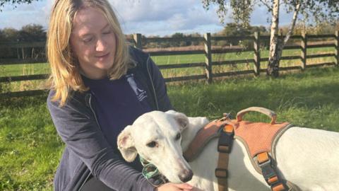 A blonde woman is crouching next to a white dog, petting it. She is wearing a navy hoodie over a navy t-shirt, with her company logo on it. She is looking down at the dog, who is wearing an orange harness. They're in a field, with a fence and trees behind them.