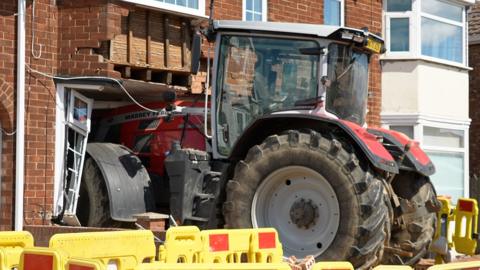 A red tractor has gone into the front of a semi-detached house, caving in the window and some of the brickwork. At its height, the tractor reaches the top window. The scene is surrounded by yellow and red temporary barriers.