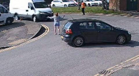 A black car and a woman with a white top with her hands out to her side in front of a white van at a t-junction