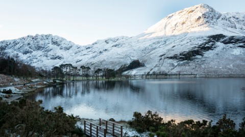Snow on the hills around a lake called Buttermere in the Lake District, Cumbria. The sun glistens on the water. The picture was taken in 2019.