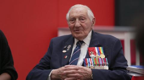 a world war two veteran sits with his hands resting together wearing lots of medals and badges on a smart suit