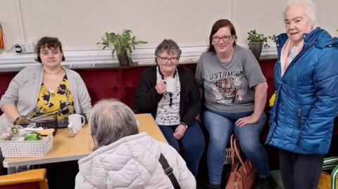 Four women sitting in the warm space smiling with mugs of tea.