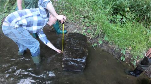 A man in blue checkered shirt, jeans and welly boots is leaning over a large stone block on the edges of the river as he measures the block. The boots of two other people looking on are also visible.