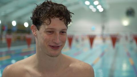 A man with short dark hair is topless, standing in front of a swimming pool.