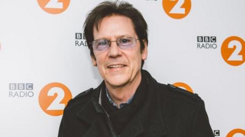 Shakin' Stevens pictured in 2021. He has short dark hair and is wearing glasses. He is smiling at the camera and is wearing a black jacket. He is pictured from the chest up, in front of a ˿ Radio 2 billboard.