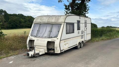 A dirty caravan parked in a country lane with a hedge running alongside and fields in the background