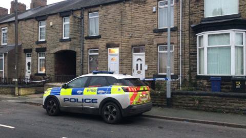 A police car is parked outside a row of terraced houses. One of the houses has blue and white police tape strung outside it. 