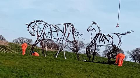 Three workman install two large horst statues. The statues are made of steel and are designed to look as though they have been drawn with pens.
