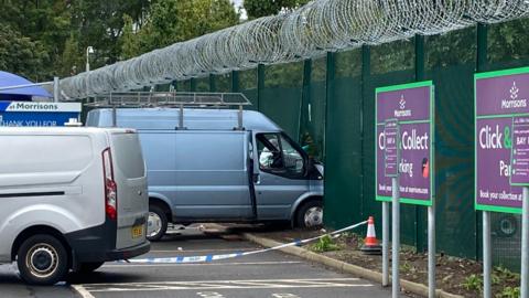 A van which has been driven into a fence in Edinburgh. The van is light grey in colour. It has a silver roof rack. The front has buckled the fence, which is green and topped with barbed wire.
