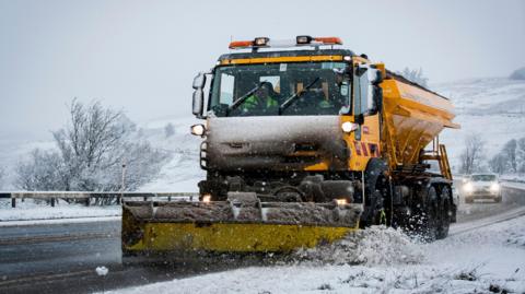 A yellow road gritter with a plough on the front driving down a rural road. The road is surrounded by a snow-covered landscape. Two cars can be seen on the road behind the gritter.