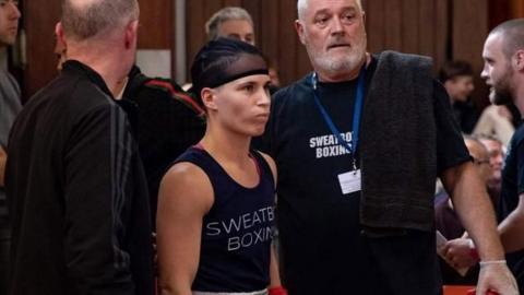 A female boxer wearing a cap and blue shirt has her cornerman by her side with a black towel over his shoulder. They are both in a crowded room.