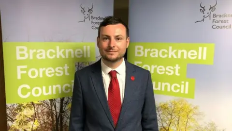 Labour's Peter Swallow wearing a dark grey suite and red tie and pin badge. He has short dark hair and short moustache and beard. He is standing in front of two Bracknell Forest Council banners with the council logo in green on them as well as a photograph of frees in the sunlight