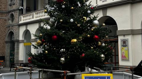 The bottom of a decorated Christmas tree on Market Street in St Peter Port, Guernsey. There is gold tinsel wrapped around with red and white baubles. There is a gold star at the top and buildings in the background 