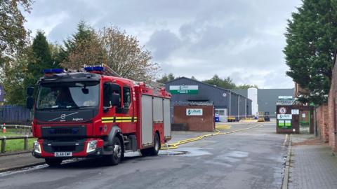 A red fire truck with yellow hoses trailing out the back leading to a factory 