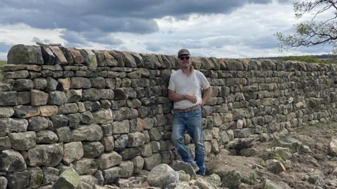 A man wearing a grey polo shirt, blue jeans and a green cap leans against a dry stone wall, which is about the same height as he is. 