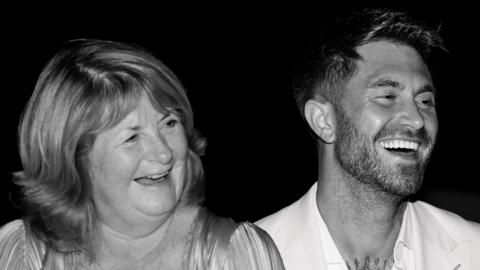 Ross Worswick and his mother Judith are sitting side by side and are laughing while staring off camera in this black and white photograph.