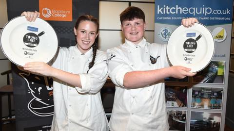 Connie and Noah in chef's whites, holding up plates that say "Young Chef" in a black graphic that looks like a frying pan. Connie has dark hair in a plait and Noah has short dark hair, both are smiling
