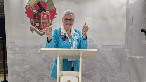 Anita Stanley wearing a light blue suit with her thumbs up and smiling at the camera