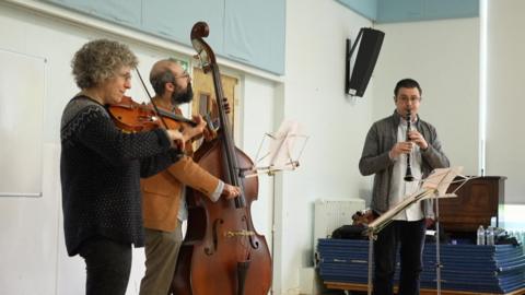 Three people in a band. One woman plying a violin, a bearded man playing a double bass, and a man playing a clarinet