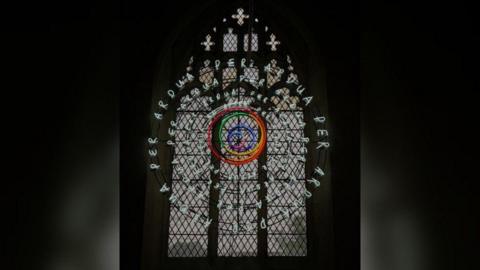 The white neon words "Per Ardua" against a large arched church window in a spiral. In the middle are the Pride colours in a spiral.