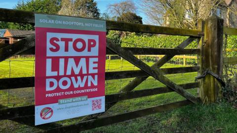 A red sign with white writing urging people to "Stop Lime Down" is fixed on a five bar wooden gate. Behind the gate is a grassy field, and a stone house.