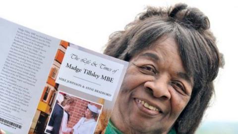 Madge Tillsley with dark hair is smiling at the camera. She is holding up, level with her face, a publication called The Life & Times of Madge Tillsley MBE.