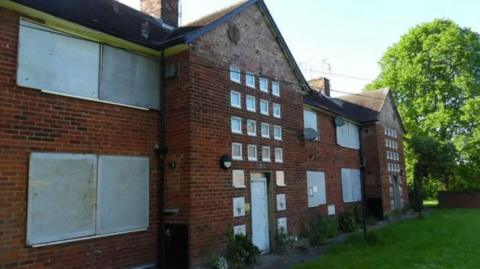 The exterior of Henson Villas in Pearson Park. It is a red brick building with multiple large windows boarded up, as well as multiple small windows. There is grass in front and green trees can be seen in the background.