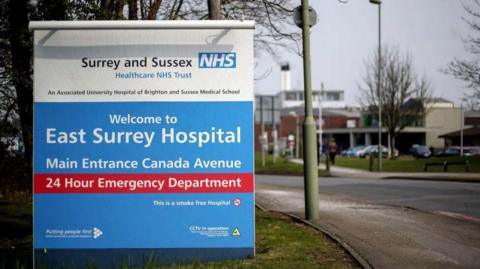 A sign blue outside the East Surrey Hospital, with Surrey and Sussex Healthcare NHS Trust badge on it
