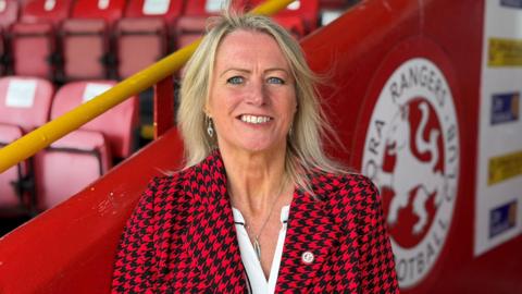 Mary Stewart has blonde hair and is wearing a red and black patterned jacket over a white top. She is smiling as she stands outside at Brora Rangers' ground.