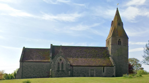 A church on a grass hill. Its tower on the right is slightly tilted. The sky is blue.