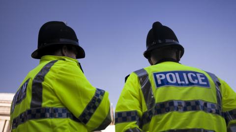 Image of two police officers in florescent jackets. POLICE is seen on the back and they're wearing black police hats. The sky is blue above them. 
