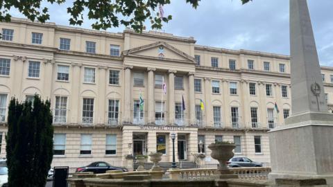 The exterior of Municipal Offices in Cheltenham, a large regency building with flags outside.