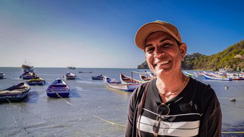 Fisherwoman Yurmari Martínez smiles a she poses for a photo. She is wearing a yellow baseball cap and a black-and-white stripy sweater. Her glasses are tucked into the collar of her sweater. Behind her, small fishing boats float in a bay. Th