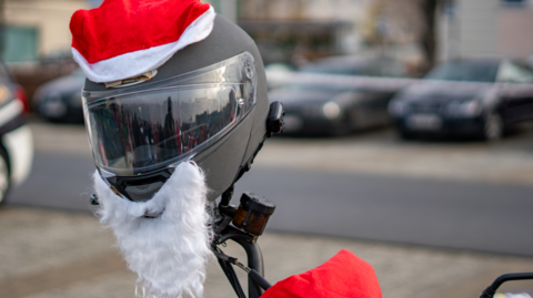 A helmet is placed on a motorbike's handlebars, dressed with a santa hat and beard.