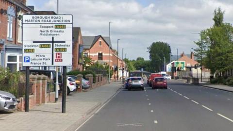A streetview of Borough Road in Middlesbrough. There are terraced houses on the left with a large sign reading Borough Road/ Marton Road Junction. Cars are parked at traffic lights down the street.