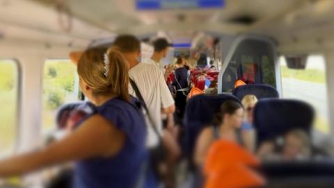 A photograph taken looking down one of the packed carriages on the train.  The seats are full and people are standing all the way down the middle.  The image is blurred so people's faces cannot be seen.