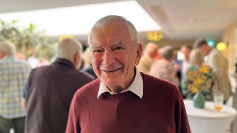 Major Bob Place is a man in his man in his 70s-80s. He has white hair and eyebrows and his smiling showing teeth. He has happy blue eyes.  He is wearing a burgundy cashmere-looking jumper and his white checked shirt is poking through at the collar.  