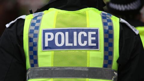 The back of a police vest. It is neon yellow and has two vertical stripes in navy and grey chequers. A Navy blue rectangle says "police" on in white lettering.