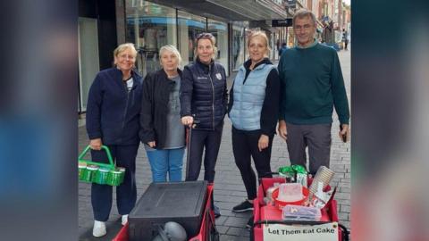 Four Let Them Eat Cake volunteers on Lincoln high street looking at the camera with two trolley bags of food and hot drinks to give out to people