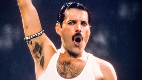 Freddie Mercury wearing a white vest and singing with his right arm aloft