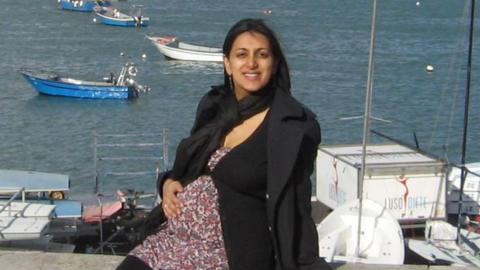 Geeta is heavily pregnant, sat outdoors with the coast behind her and small boats on the water. She is wearing a floral long top, with black trousers, cardigan and jacket and has her hand on her bump.