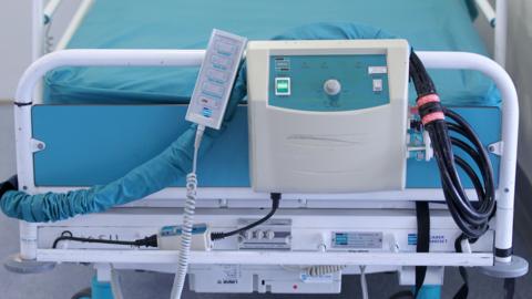 A stock image of a hospital bed. There is a metal bed frame and blue sheets/mattress within the hospital room.