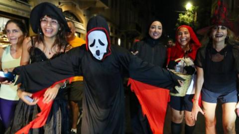 Six girls in halloween costumes, one dressed as a ghost wearing the scream mask